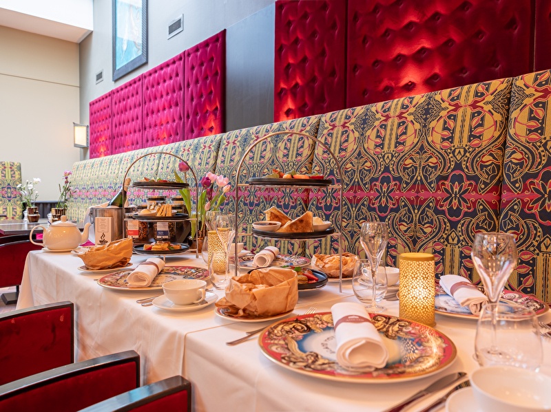 Atmospherically set table for a luxurious high tea at Grand Hotel Amrâth Amsterdam, with colorful crockery, stands filled with sweet and savory treats, teapots and elegant glasses in a stylish and warm setting.