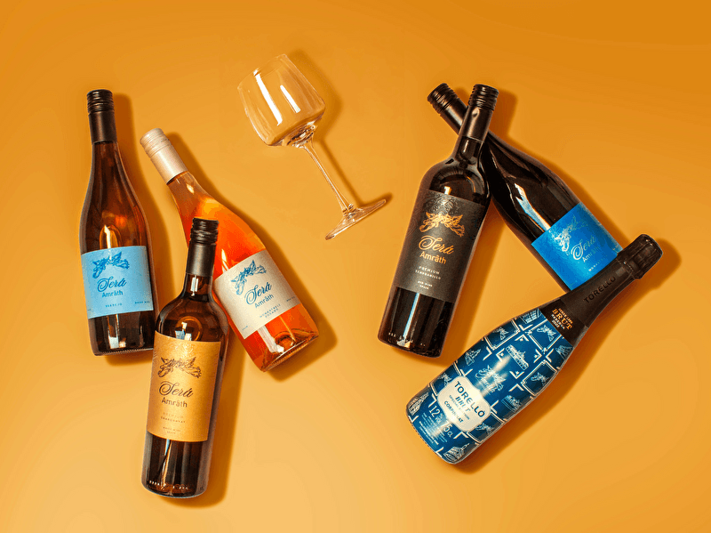 Vijf Será Amrâth-wijnen en één Torelló-fles, samen de Amrâth Exclusives wijnselectie, op een oranje achtergrond. De flessen hebben diverse etiketten in blauw, zwart, wit, oranje en rosé. Een leeg wijnglas ligt in het midden.