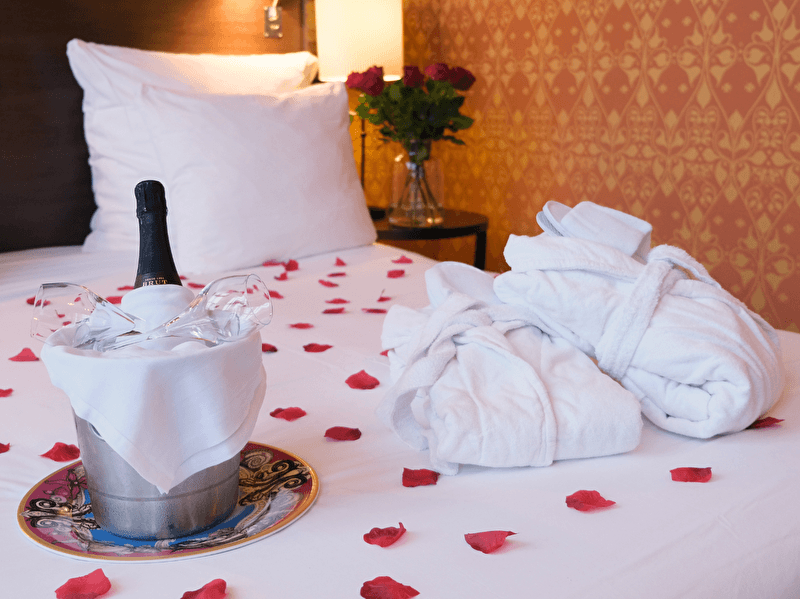 A romantically prepared bed with rose petals, a chilled bottle of champagne in an ice bucket, two glasses, and two white bathrobes. In the background, a vase with red roses is visible.